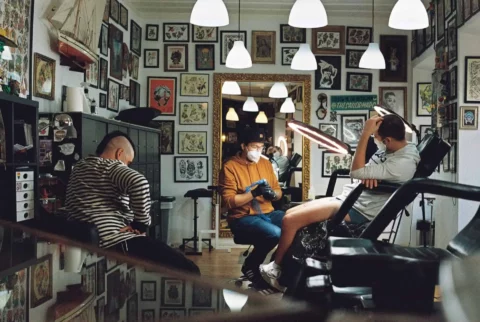 Three men sitting in a tattoo shop. One man is preparing a tattoo machine and another is preparing to be tattooed