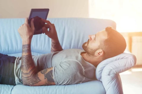 man with arm tattoos laying on a couch holding an iPad
